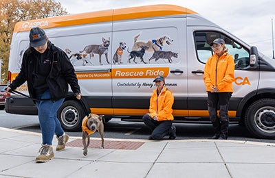 A dog with three legs is led away from an ASPCA Rescue Ride van by an ASPCA team member