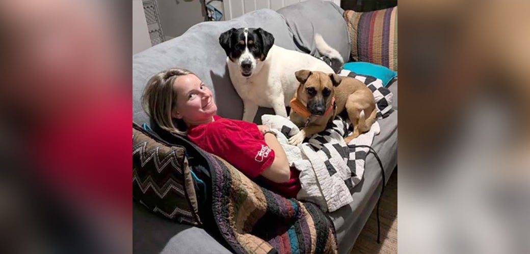 A woman in red shirt on a couch with two dogs laying on her