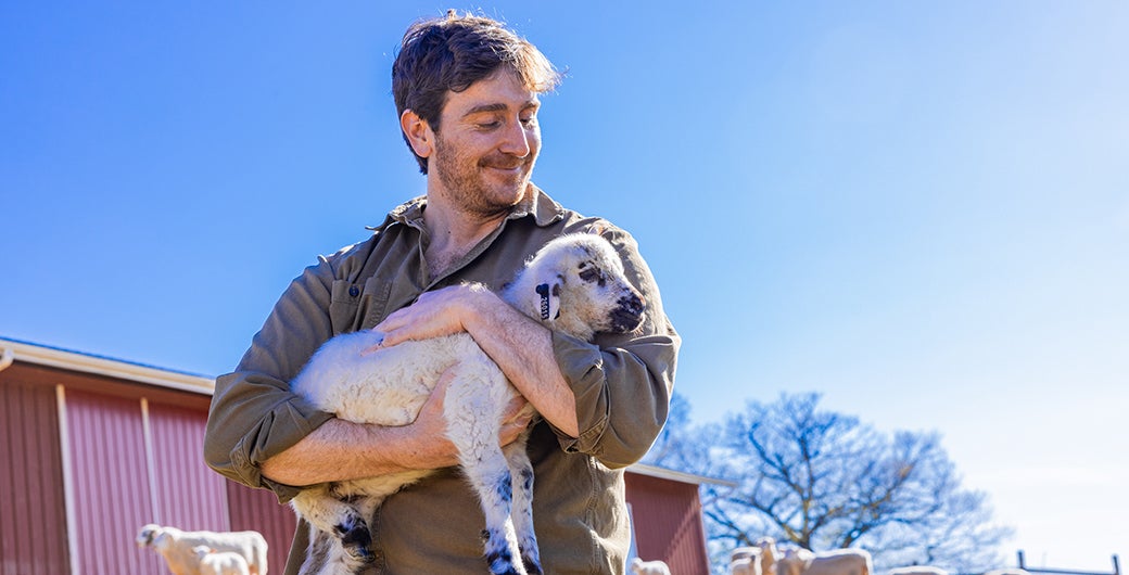 Farmer smiling while holding a baby goat with other goats nearby grazing on pasture.