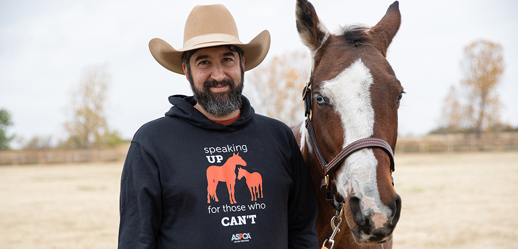 A man with a cowboy hat, salt and pepper beard, and an aspca horse hoodie next to a brown and white horse